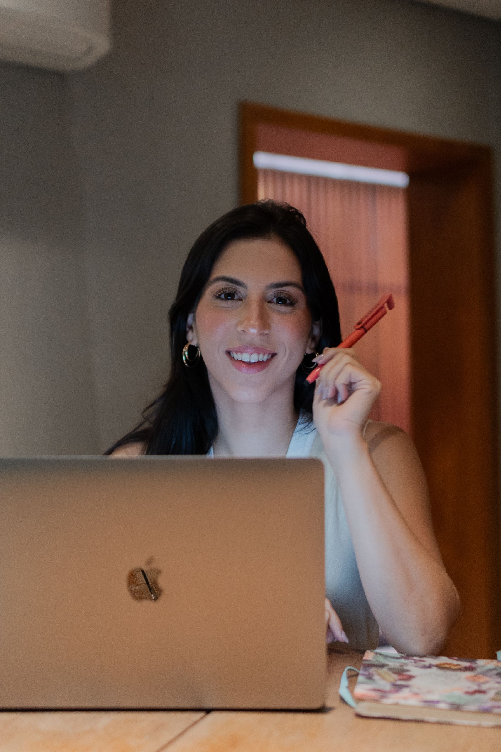 Smiling woman holding pen while working on laptop at desk.